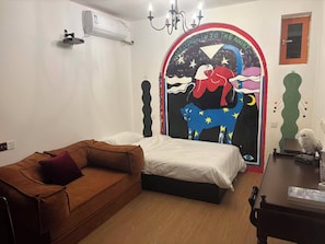 A bedroom with a bed, a brown sofa, an air conditioning unit, a chandelier, and a wall with a colorful mural.