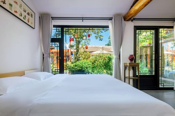 A hotel room with a bed, a window with curtains, a small table with a vase, and a view of greenery outside.
