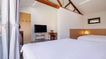 A hotel room with a large bed, a desk with a flat-screen TV, a bedside table with a lamp, and a wooden ceiling beam.