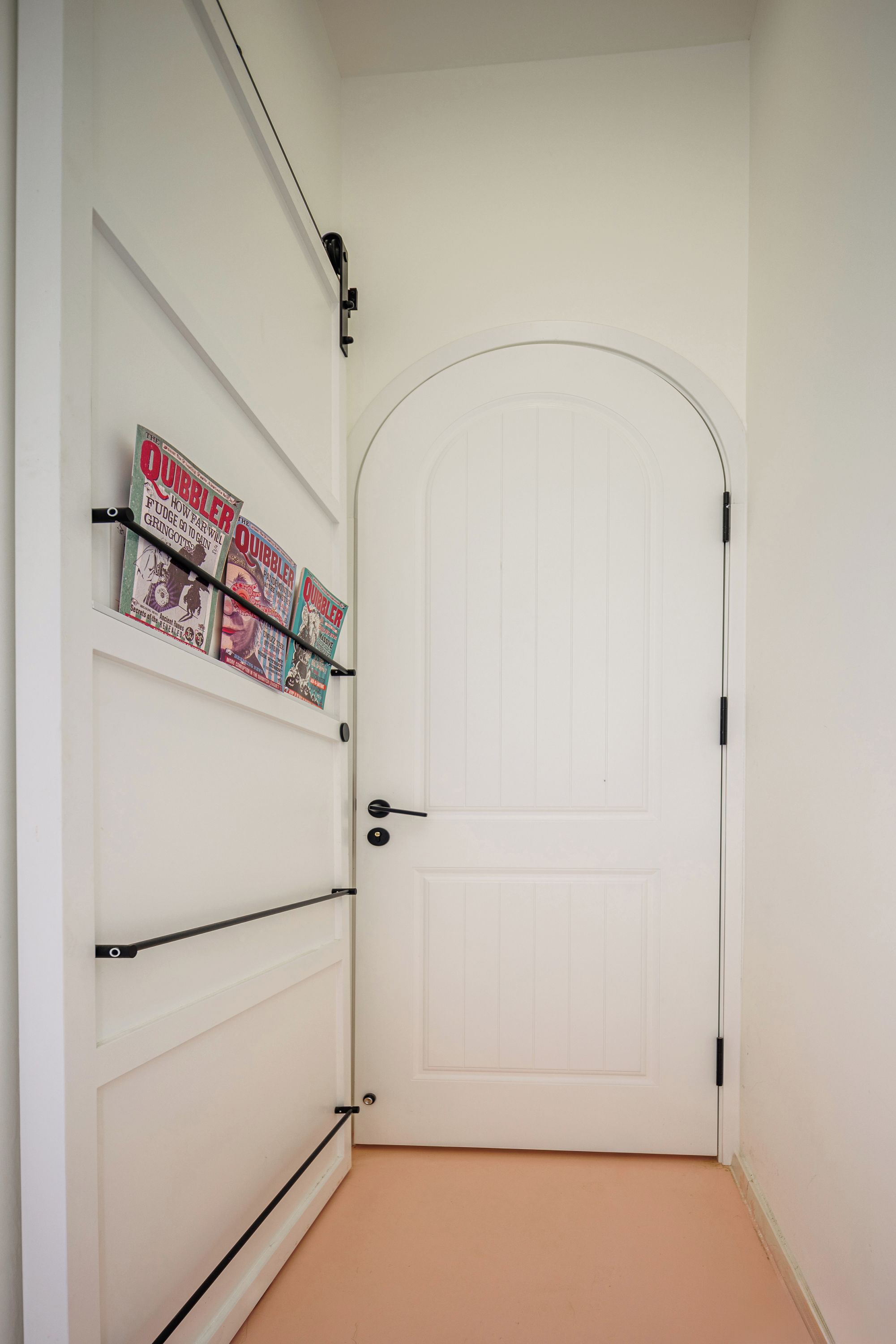 A white door with a black handle, a white wall with a shelf holding several Quibbler magazines, and a black cable running along the wall.