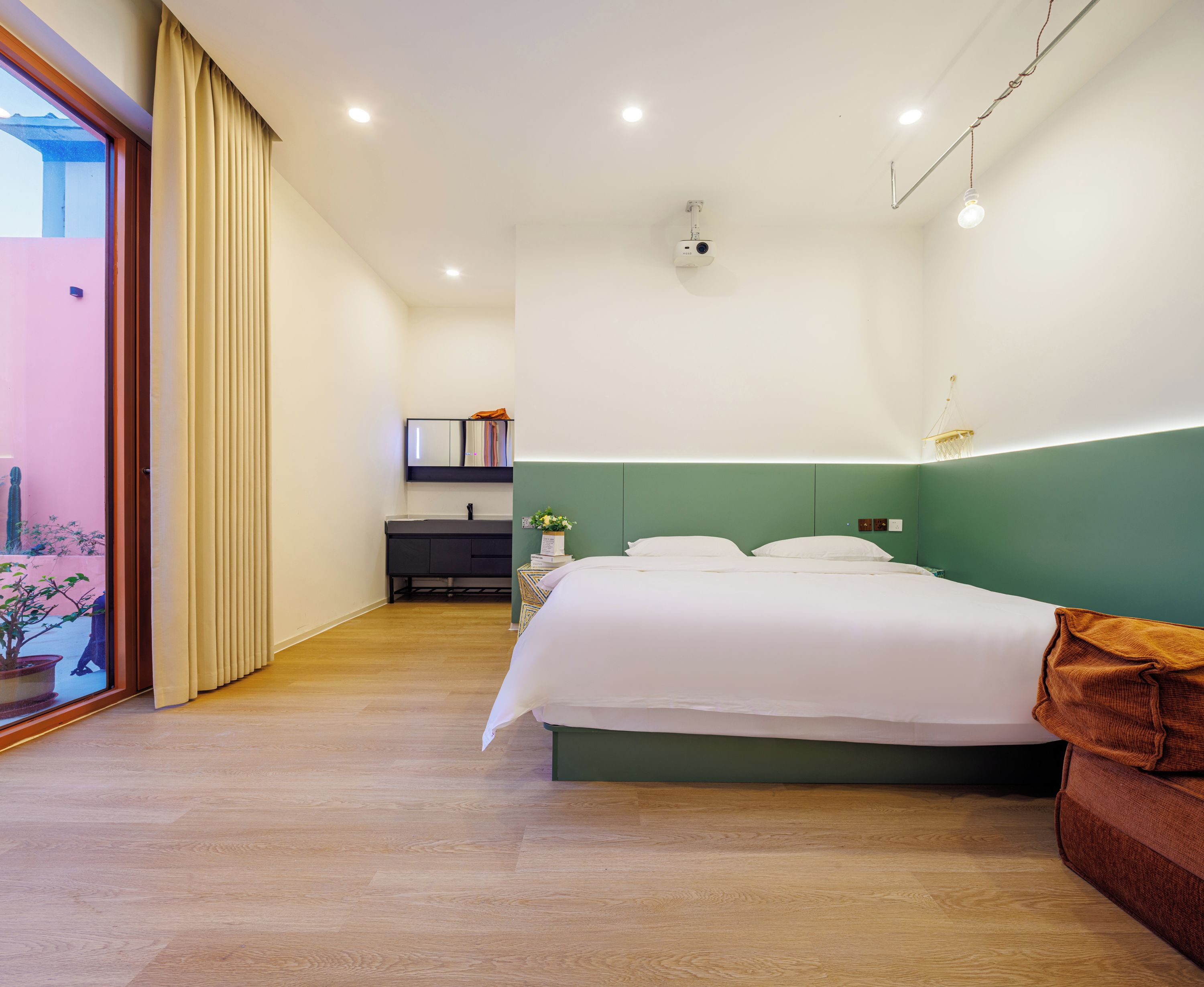 A hotel room with a bed, a bedside table, a mirror, a green accent wall, and a view through a sliding door.