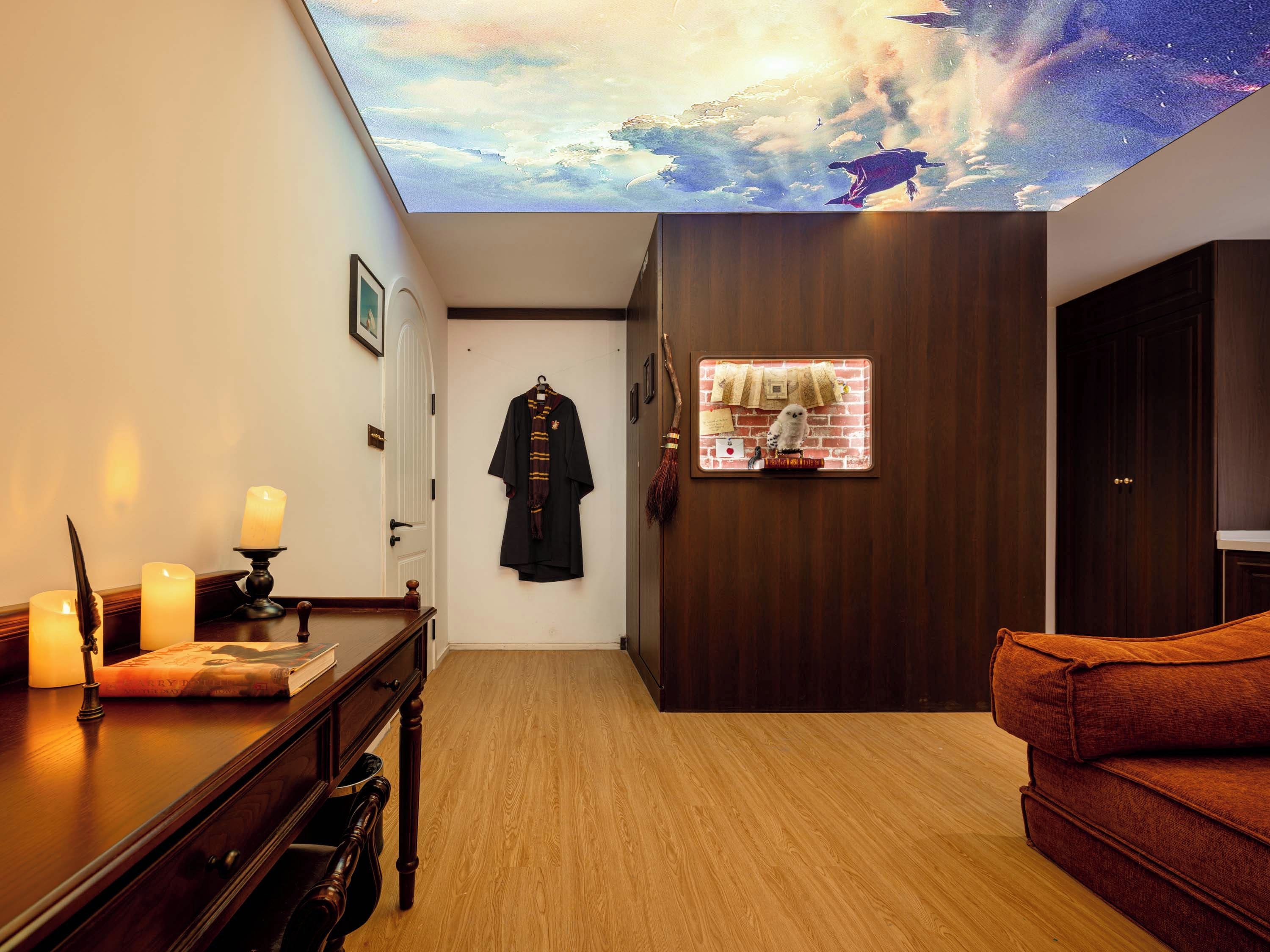 Una habitación con piso de madera, un escritorio con un libro y velas, una bata colgada en la pared y un mural en el techo.