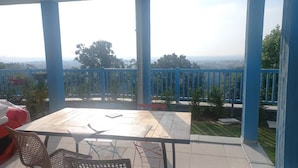 Un espacio al aire libre techado con una mesa y sillas, vista al paisaje y barandilla azul.