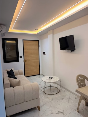 Un saló modern amb un sofà, una taula de centre rodona, un televisor fixat a la paret i una porta de fusta.
