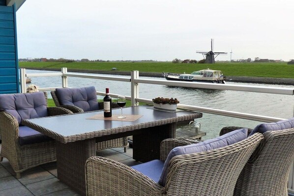 An outdoor seating area with wicker furniture and a table, overlooking a canal with a windmill and a boat in the background. - Friesland Zilverreiger 4 (JK Workum)
