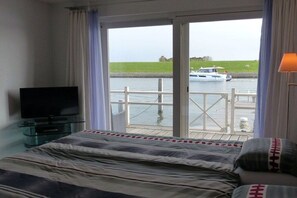 A hotel room with a bed, a television on a glass table, and a view of a canal with a boat. - Friesland Zilverreiger 4 (JK Workum)