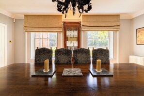 Ein Esszimmer mit einem großen Holztisch, drei Stühlen mit gemusterten Sitzbezügen, einer Chandelier und einem Fenster mit Blick auf Grünflächen.