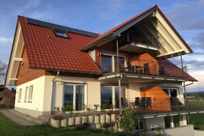 Ein zweistöckiges Haus mit rotem Ziegeldach, einem Balkon im oberen Geschoss und einem Eingangsbereich mit Glastüren.
