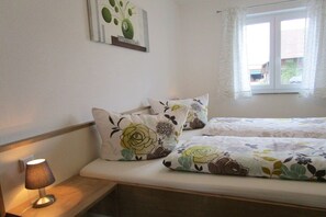 Un dormitorio con cama, dos almohadas, una lámpara de noche, una ventana con cortinas e imagen colgada en la pared.