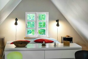 A room with a sloped ceiling, a bed with red pillows, a window with a view of greenery, and two wall-mounted lamps. - Thatched timber-framed house by meadow and stream (Balle)