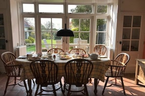 A dining area with a table set for six, surrounded by chairs, with a view of a garden through large windows. - Holiday home de Broodkiste (Domburg)
