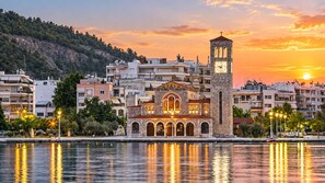 A waterfront building with a clock tower, reflecting in calm water, surrounded by residential buildings and a hilly backdrop. - Secret Garden Volos - Tranquil Private Retreat (Volos)