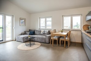 A modern living space with a grey sofa, a round dining table with chairs, a small round table with a plant, and a kitchen area with cabinets and a window. - Schottenhof Walser 3 (Achberg)