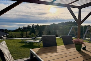 Ett takförsett utomhusområde med bord och stolar, med utsikt över ett grönområde med träd och ett staket.