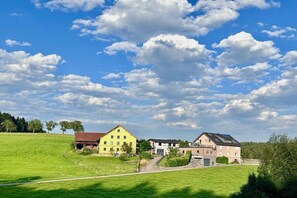 En lantlig miljö med en gul byggnad och ett modernt hus med ett sluttande tak, omgivet av gröna fält och en klar himmel.