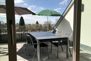 An outdoor patio with a table and chairs, a green umbrella, and a view of trees and buildings. - Holiday apartment with 1 bedroom (Lindau)