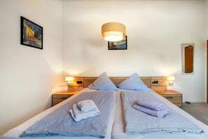 Un letto matrimoniale con due cuscini e un asciugamano piegato, un comodino con una lampada e un quadro appeso al muro.