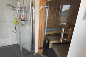 Une douche en verre avec une porte en bois, une chaise et une petite table sur laquelle sont posés des articles de toilette.