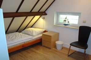 Una habitación pequeña con cama individual, techo inclinado, una ventana decorada con un velero, una mesita de noche, una silla y un cubo de basura.