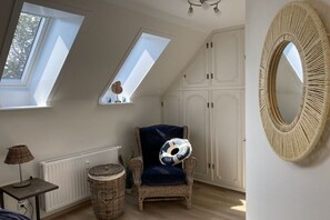 A room with a sloped ceiling, a wicker chair with a cushion, a round mirror, and a table with a lamp. - Baltic Sea Dream 139 (Travemünde)