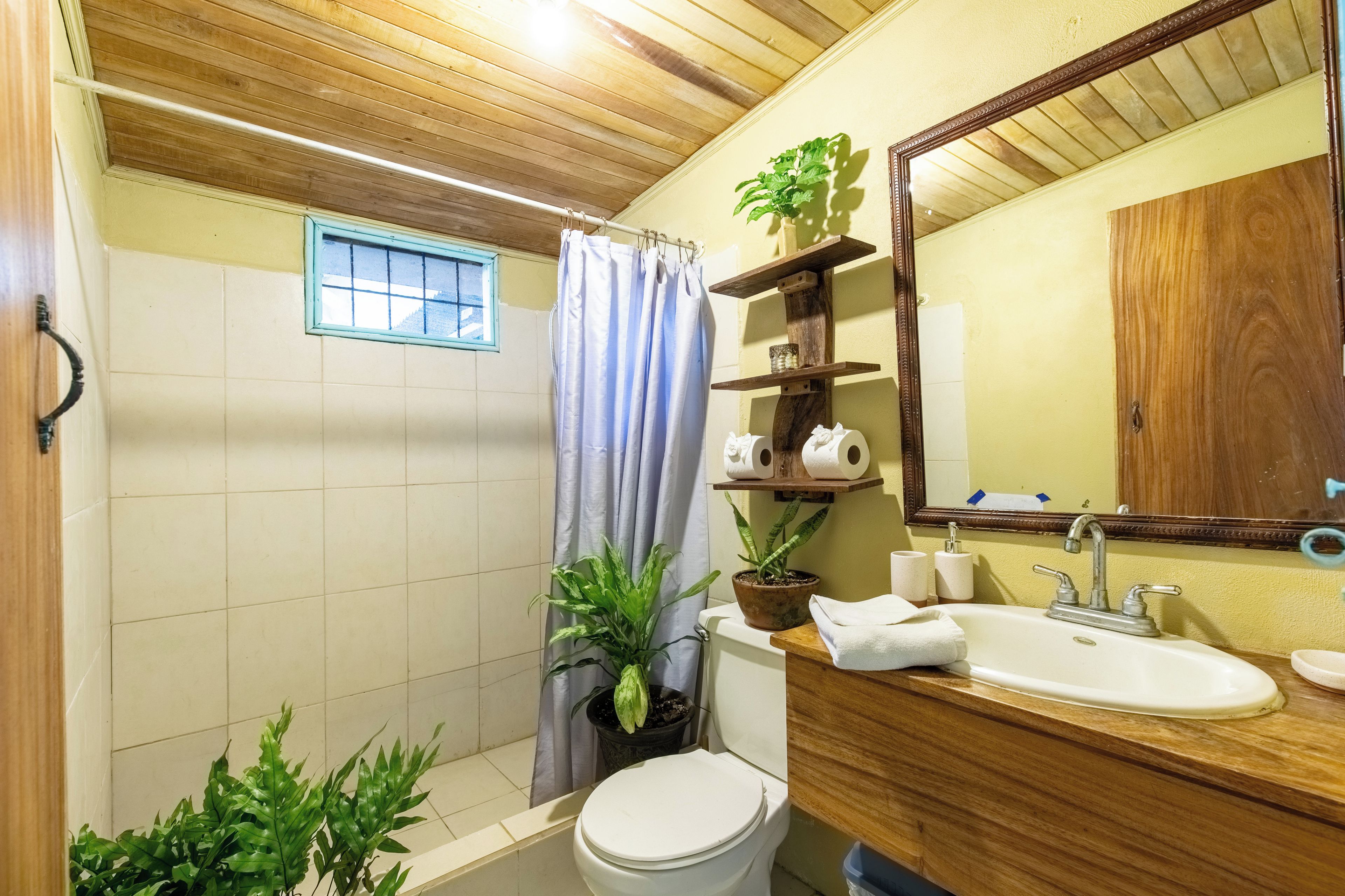 Un baño con techo de madera, inodoro, lavamanos, espejo y ventana con cortina.