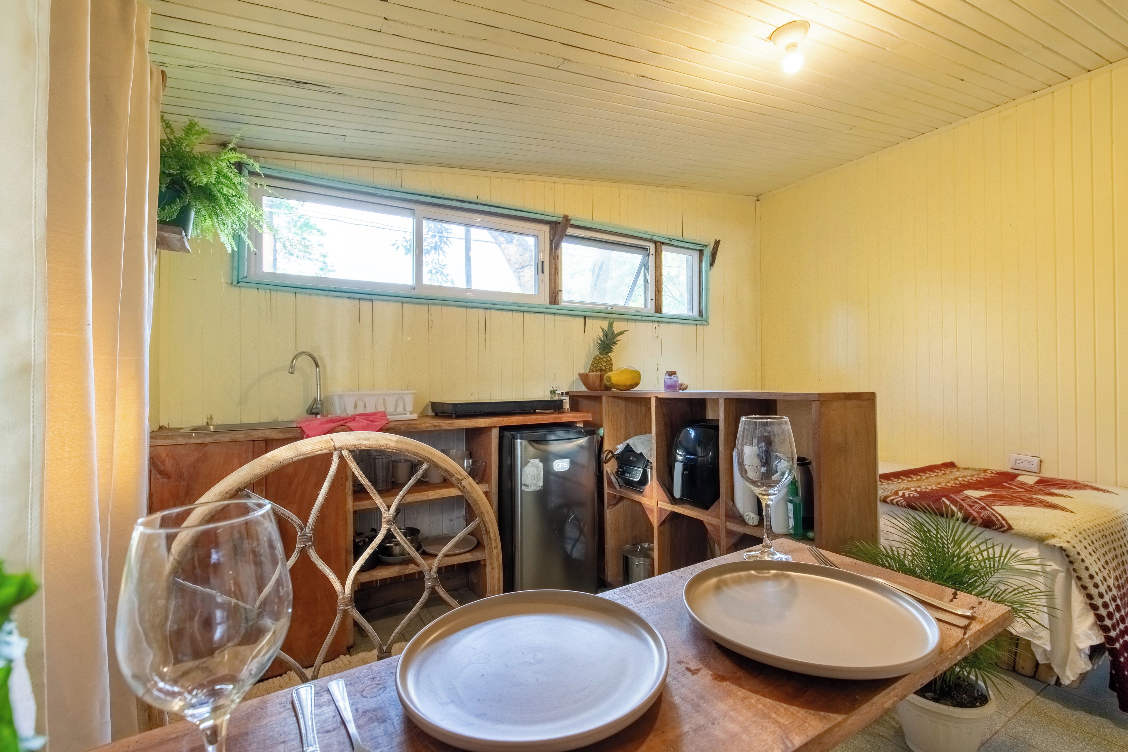 Una habitación acogedora con una mesa de madera lista para comer, una cama con manta estampada y una ventana con vistas a árboles.