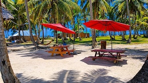A beach with two picnic tables and red umbrellas, surrounded by palm trees and a clear blue sky.