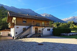 A two-story wooden house with a staircase leading to the upper floor, a paved driveway, and a mountain backdrop. - Holiday apartment with 3 bedrooms (Wald im Pinzgau)