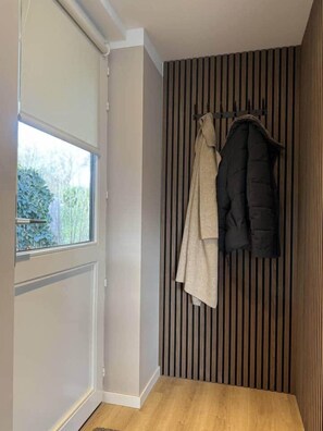 Ein Zimmer mit Holzboden, einem Fenster mit Jalousien, einem Kleiderständer mit zwei Mänteln und einer gestreiften Wand.