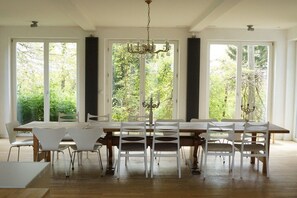 A dining room with a large table and chairs, large windows with curtains, and a chandelier.