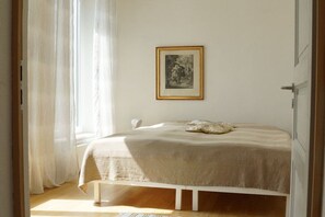 A bedroom with a bed, a framed picture on the wall, and a window with sheer curtains.