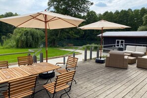 En træterrasse med udendørs møbler, herunder et bord og stole, samt to store parasoller.
