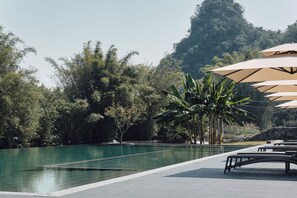 A pool area with lounge chairs and umbrellas, surrounded by trees and a mountain in the background.