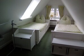 Quarto sótão pequeno e triangular com duas camas, uma janela com cortinas e um radiador.