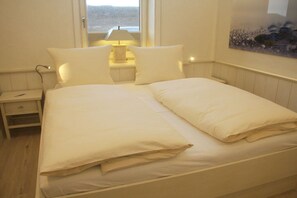 A bed with white bedding, a bedside table with a lamp, and a window with a view of the outdoors. - 1 in the Annelene house (Westerland\/ Sylt)