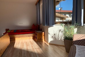 A room with a wooden floor, a red sofa, a small wooden table, a potted plant, and a view of a building with a red roof through a window with blue curtains. - Top 1 in the Alpine House (Uderns)