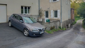 Un coche gris estacionado frente a un edificio de dos pisos con una puerta de garaje, un pequeño jardín y un camino de acceso estrecho.