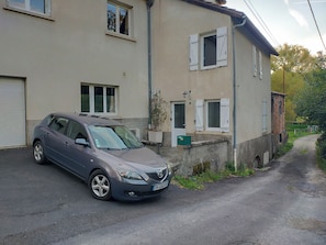 Un coche gris estacionado frente a un edificio de dos pisos con una puerta de garaje, un pequeño jardín y un camino de acceso estrecho.