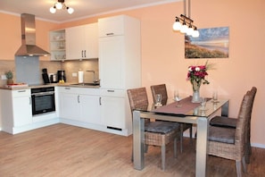 A modern kitchen with white cabinets, a stainless steel range hood, and a dining area with wicker chairs and a glass table. - Sandpipers in the Cherry Orchard 1 (Kellenhusen)