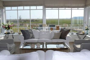 Ein geräumiges Wohnzimmer mit einem weißen Sofa, zwei Sesseln, einem Couchtisch und großen Fenstern mit Blick auf ein Feld.
