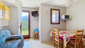 Una habitació amb un sofà, una taula amb cadires, una televisió i una finestra amb vista a vegetació.