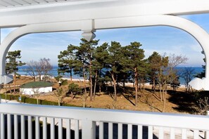 Blick von einem Balkon mit weißer Reling auf einen Strand mit Bäumen und einem klaren Himmel.