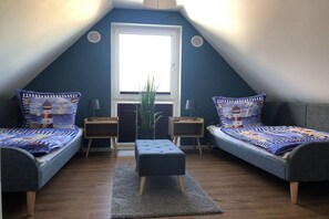 Ein kleiner, gemütlicher Raum mit schrägen Wänden, zwei Einzelbetten mit passenden Kopfteilen, einem zentralen Hocker, einer Topfpflanze und einem Fenster mit Jalousien.