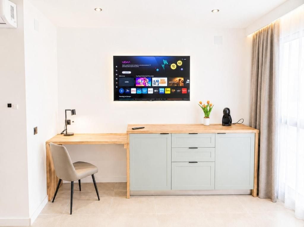 Une chambre moderne avec un bureau en bois, une armoire bleu clair, un téléviseur fixé au mur qui affiche l’interface d’un service de diffusion en continu, une chaise et une plante en pot.