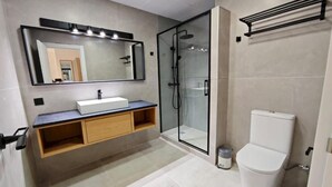 Um banheiro moderno com chuveiro em box de vidro, vaso sanitário branco e toucador com espelho e iluminação.