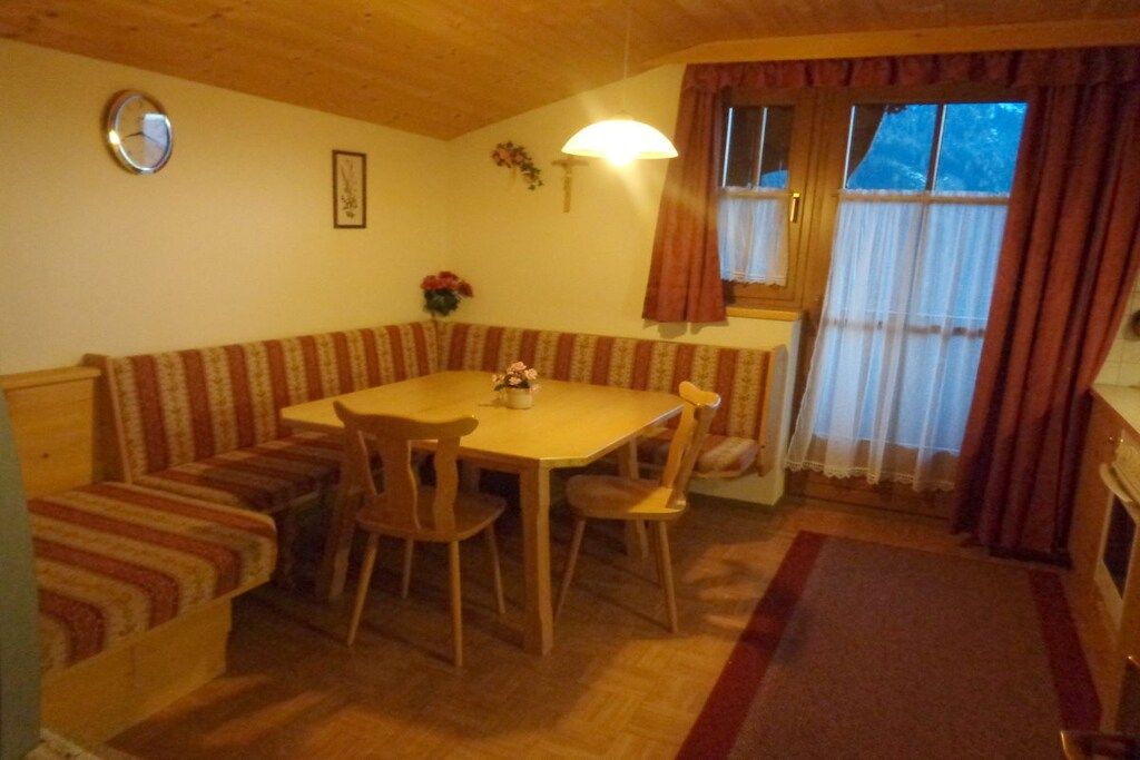 A dining area with a wooden table and chairs, a bench with striped cushions, a clock on the wall, and a window with curtains.