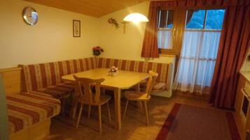 A dining area with a wooden table and chairs, a bench with striped cushions, a clock on the wall, and a window with curtains.