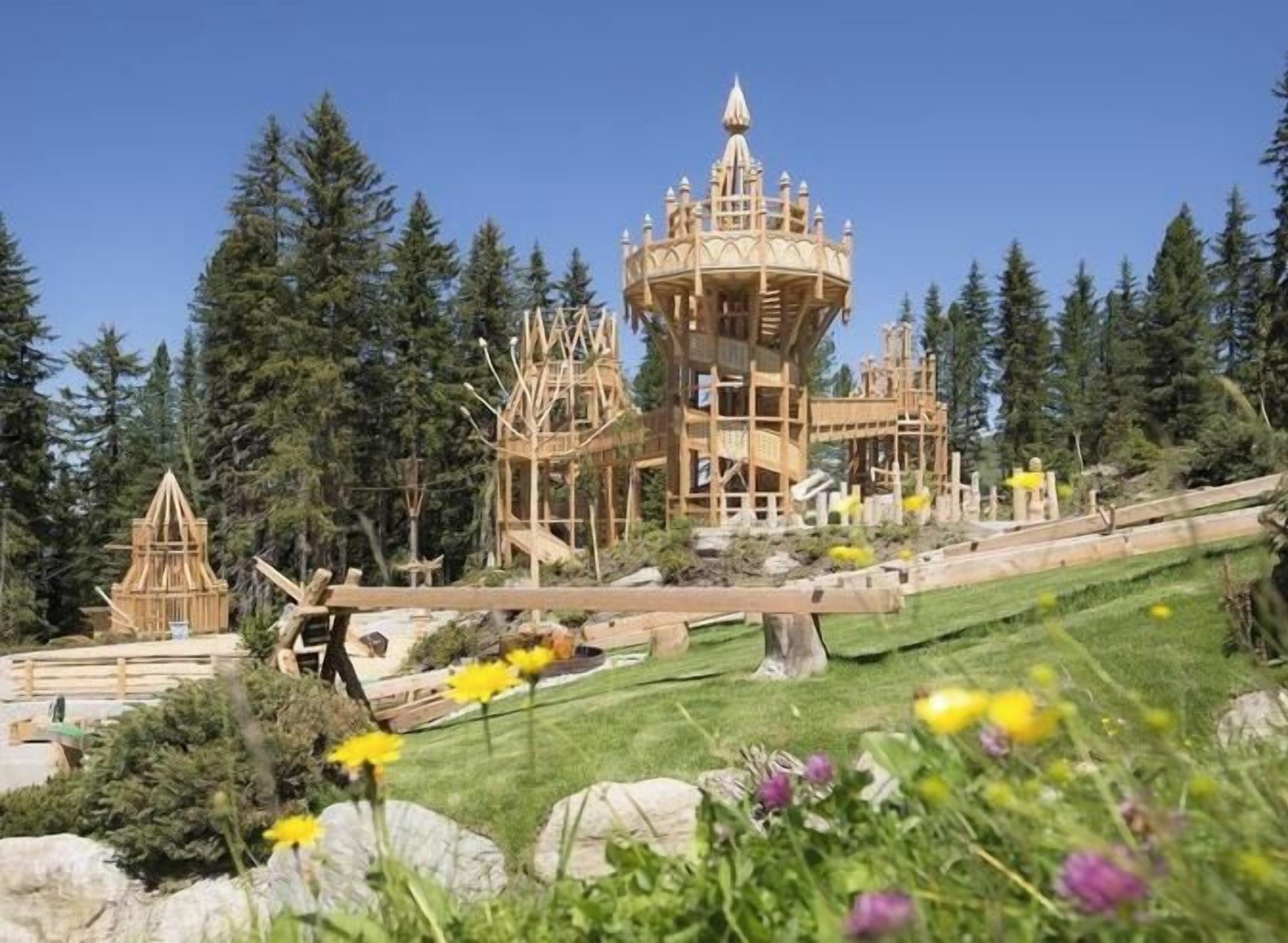A large wooden structure with multiple levels and a central tower, surrounded by a forested area with a grassy foreground.