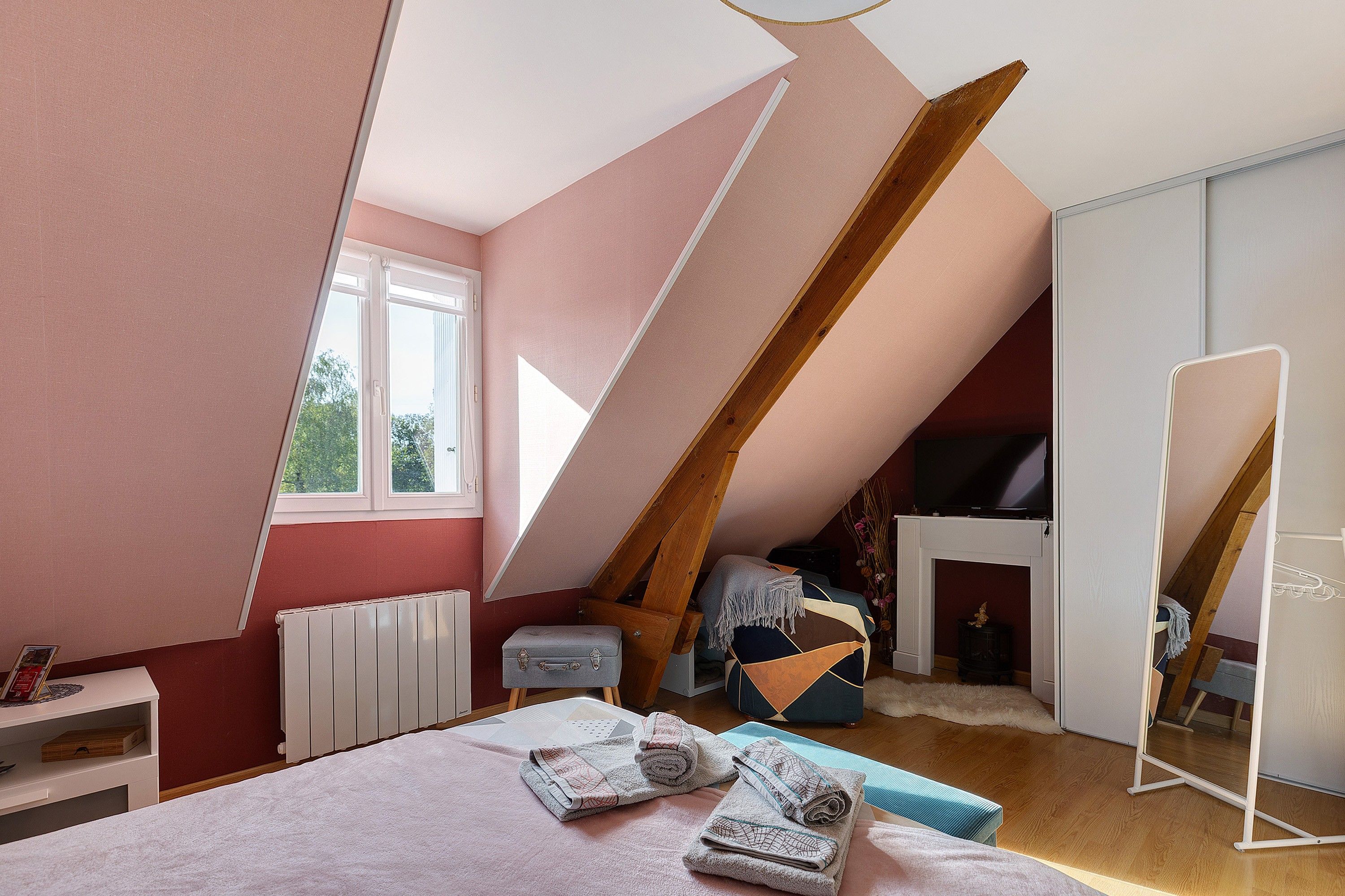 Ein Schlafzimmer mit schrägen Wänden, einem Bett, einem Heizkörper, einem Fenster, einem Spiegel und einem Fernseher.