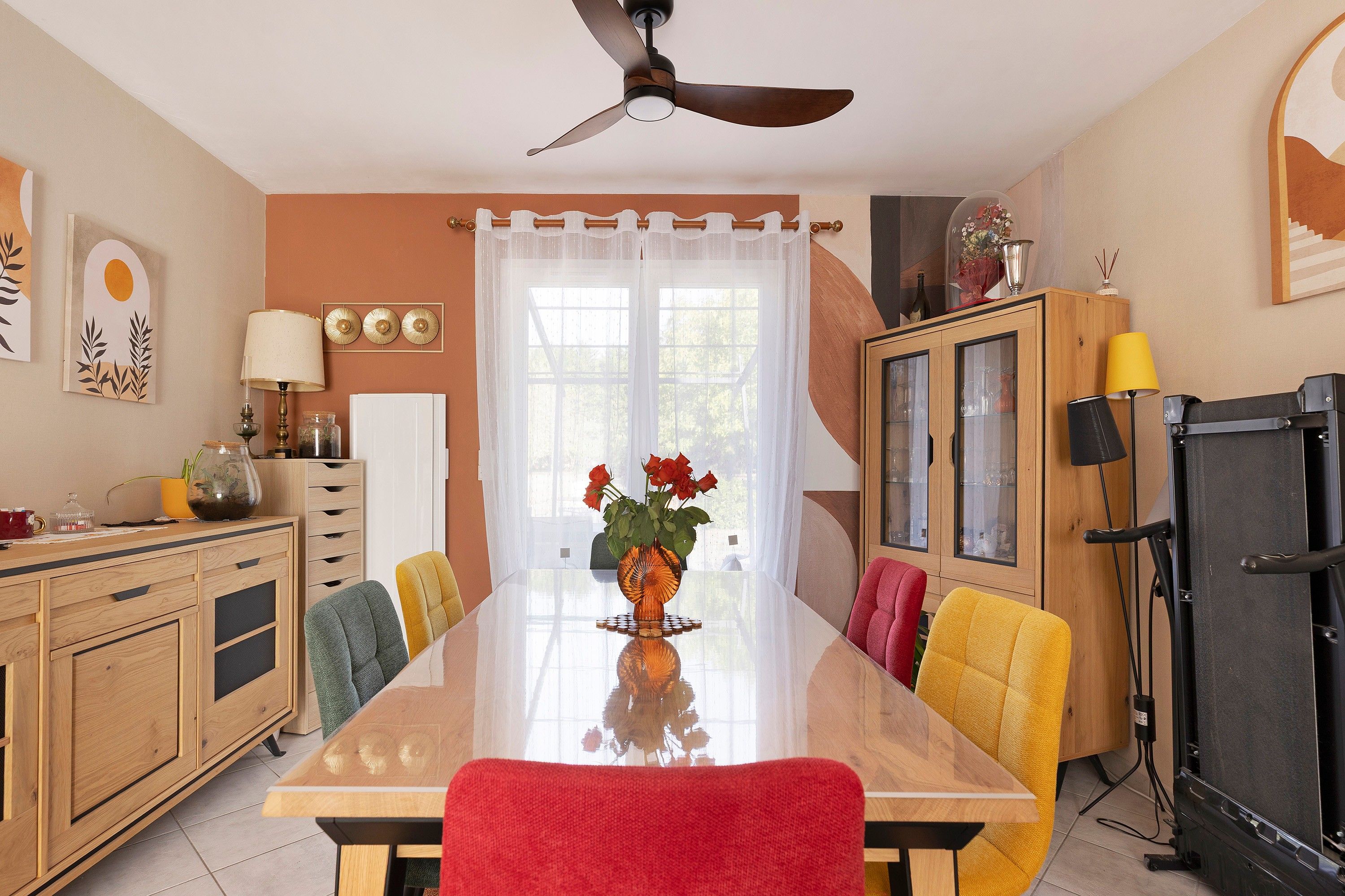 Une salle à manger avec une grande table, des chaises, un ventilateur de plafond et un buffet aux portes vitrées.